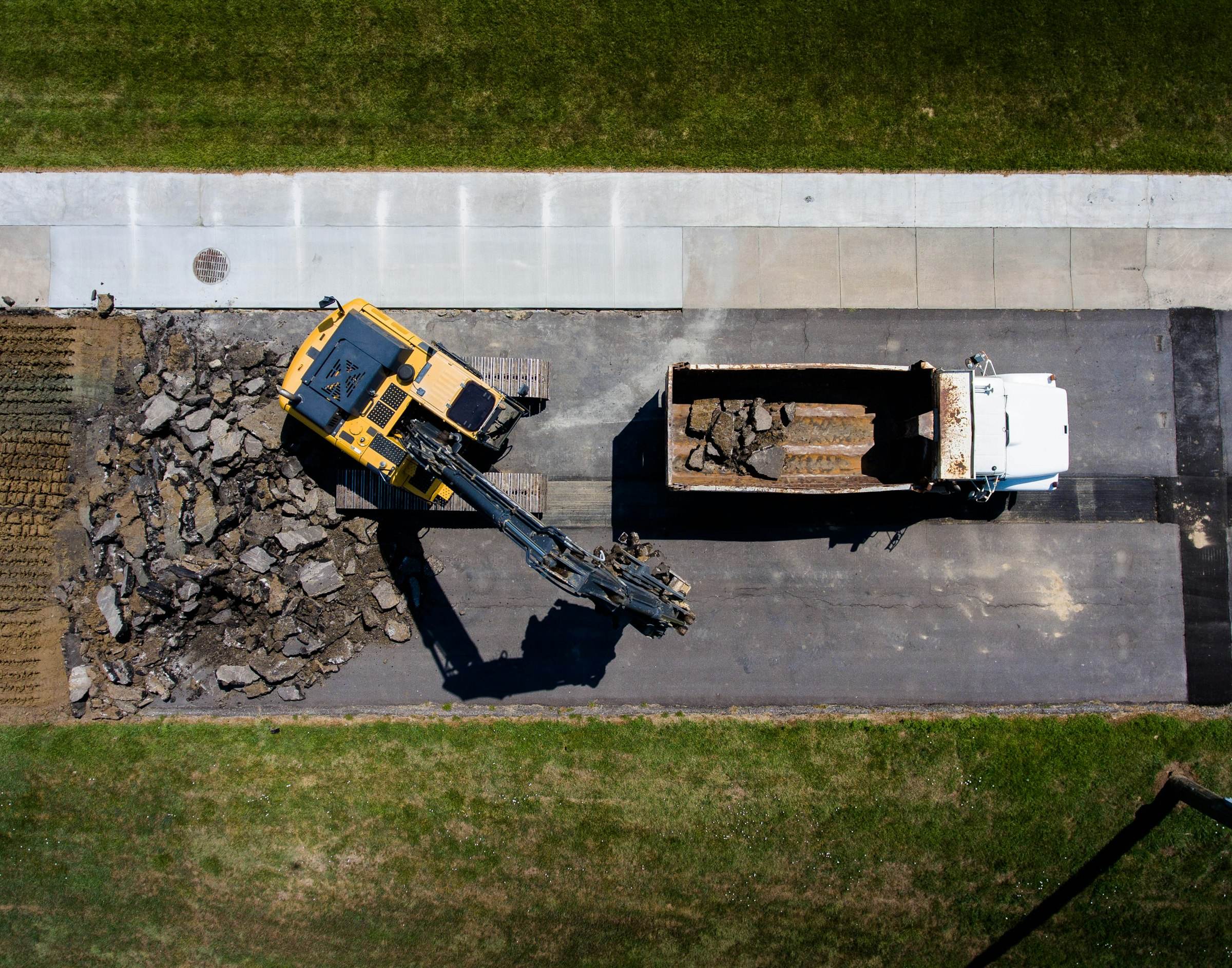 Aerial view of a yellow excavator loading broken-out concrete into a tipper lorry beside a path