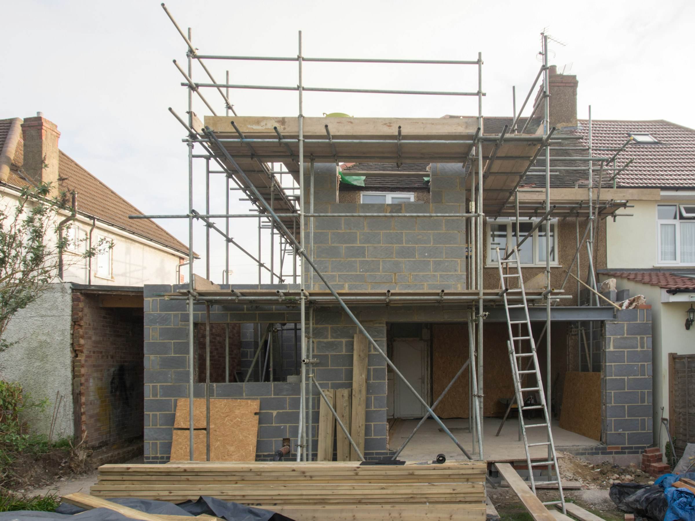 A two storey blockwork extension under scaffolding, with timber stacked in the garden